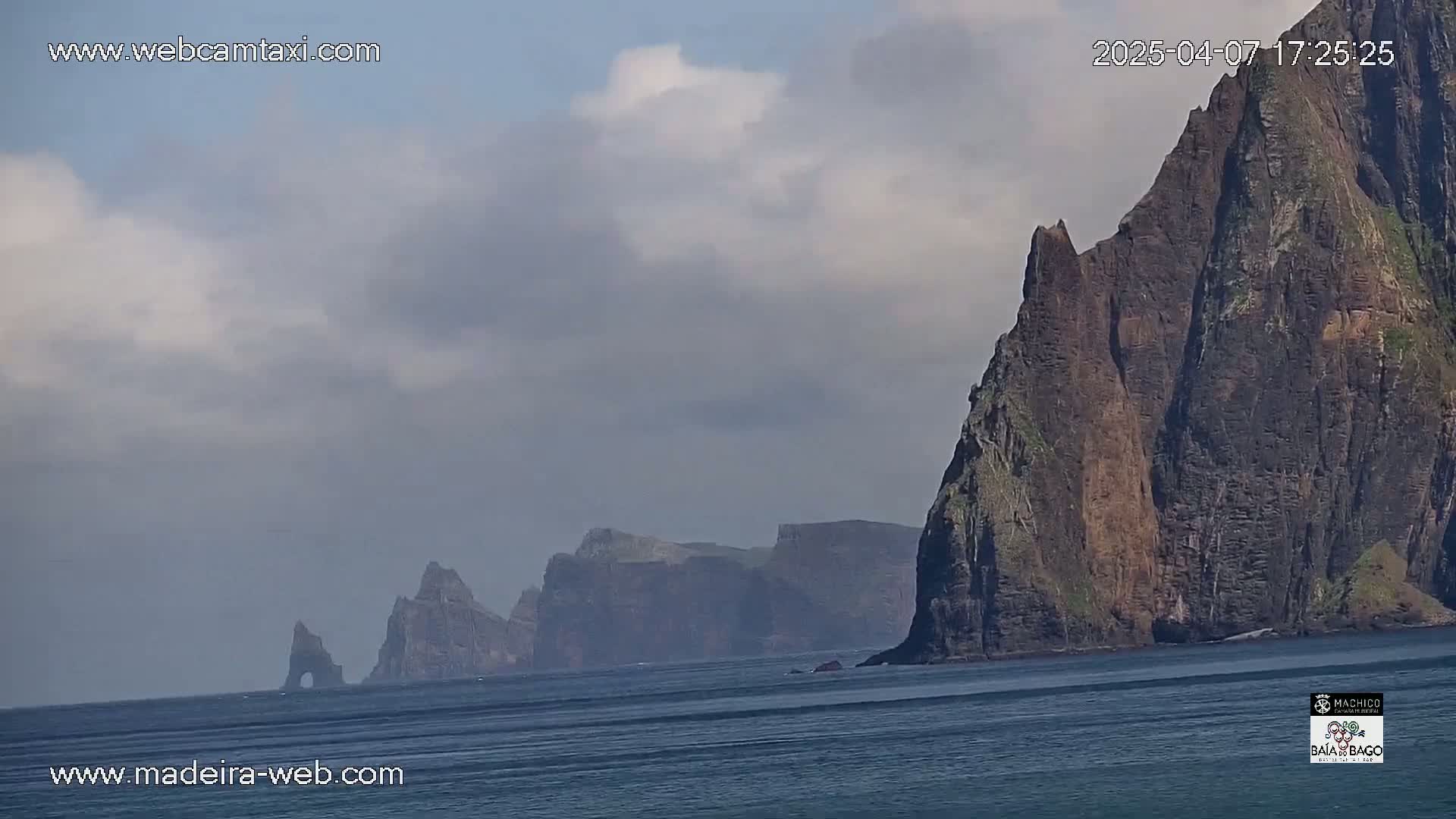 A partly cloudy day reveals a dramatic seascape featuring towering, dark cliffs rising from a calm ocean.