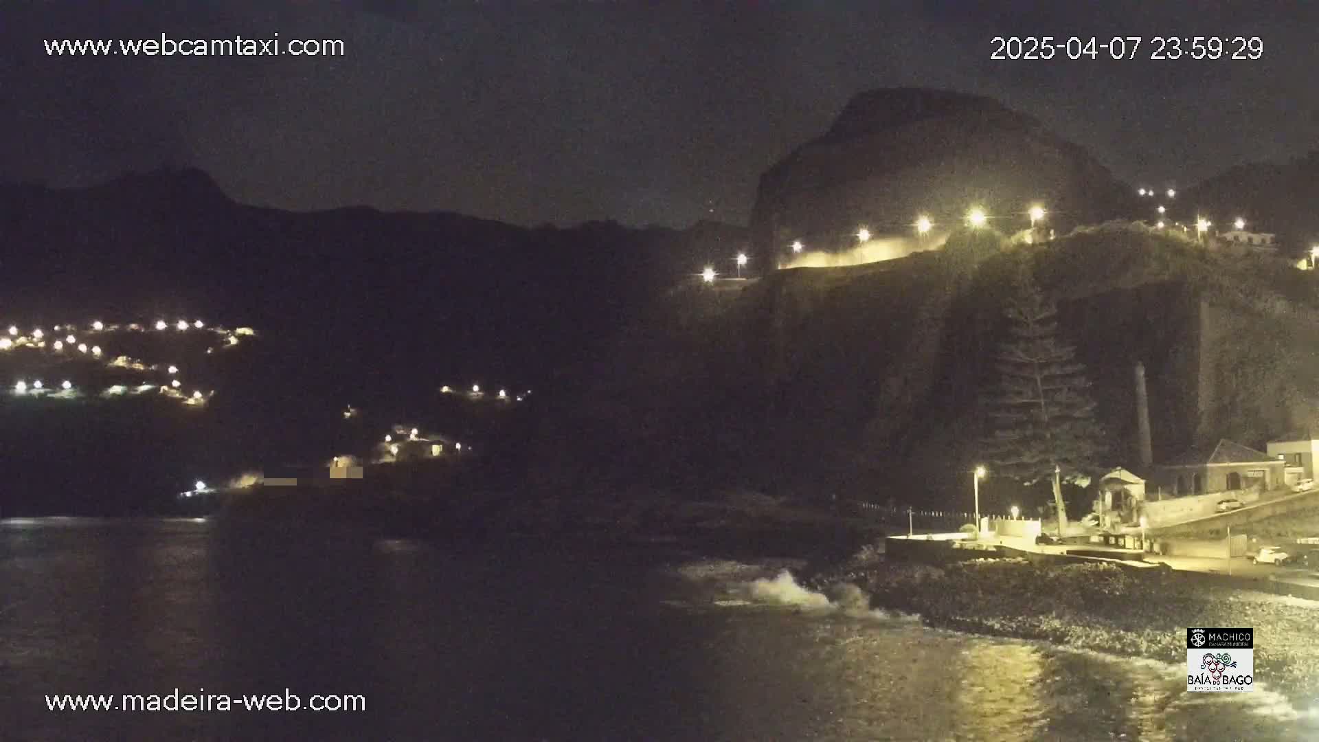 A nighttime coastal scene shows a dark, rocky cliff face with lights along a road, small buildings nestled into the hillside, and a dark ocean with small waves lapping at the shore.