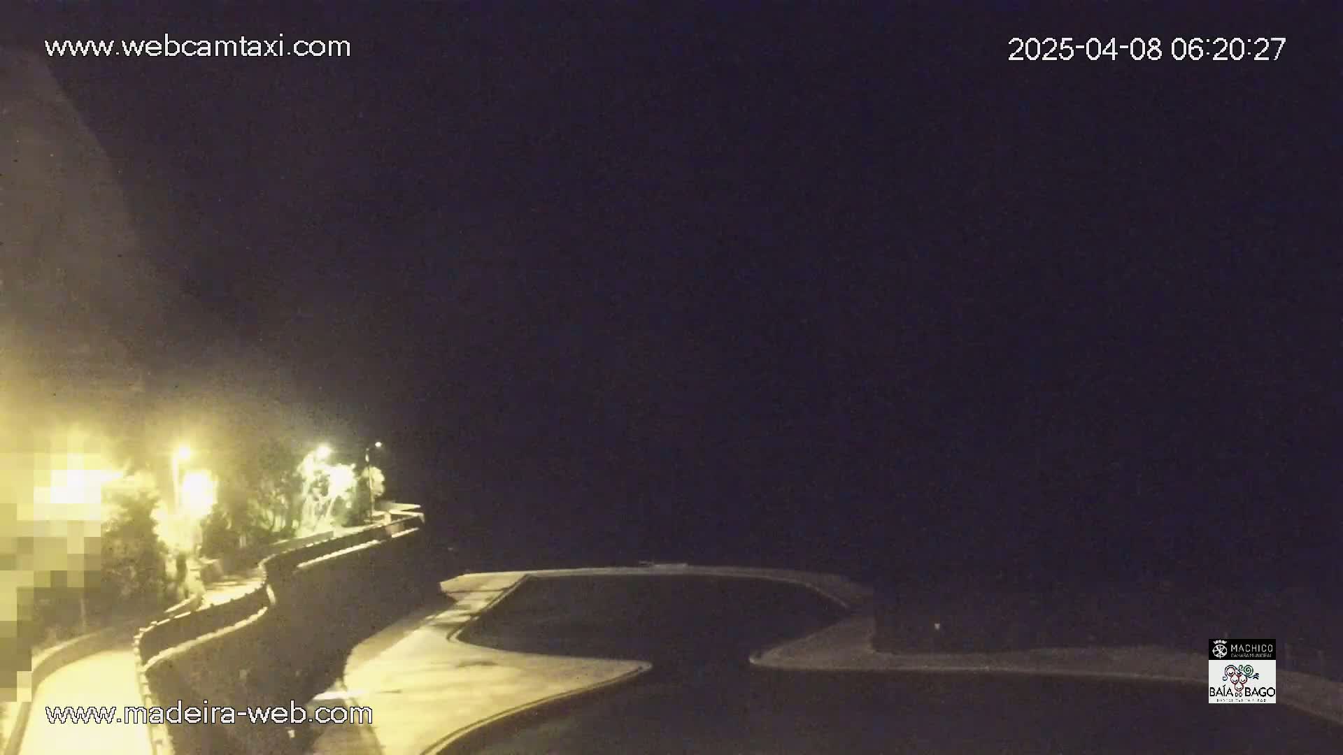 A nighttime view from a vehicle shows a roadway curving alongside a low stone wall, lined with vegetation and illuminated by streetlights.