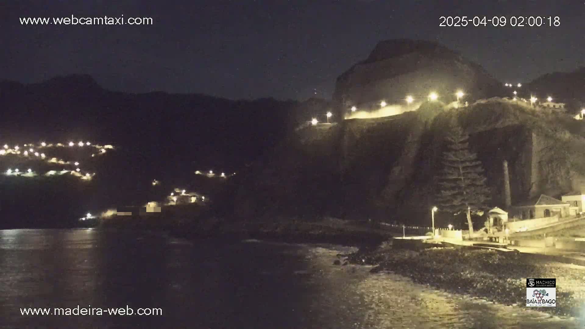 A nighttime coastal scene shows a dark cliffside with illuminated buildings and pathways, overlooking a calm body of water.
