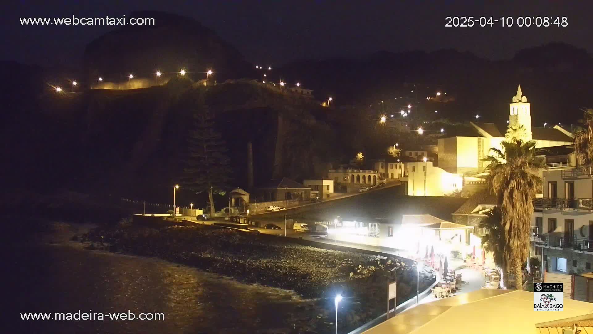 A nighttime coastal town scene, illuminated by streetlights, shows buildings on a hillside overlooking a dark, rocky shoreline with calm water.