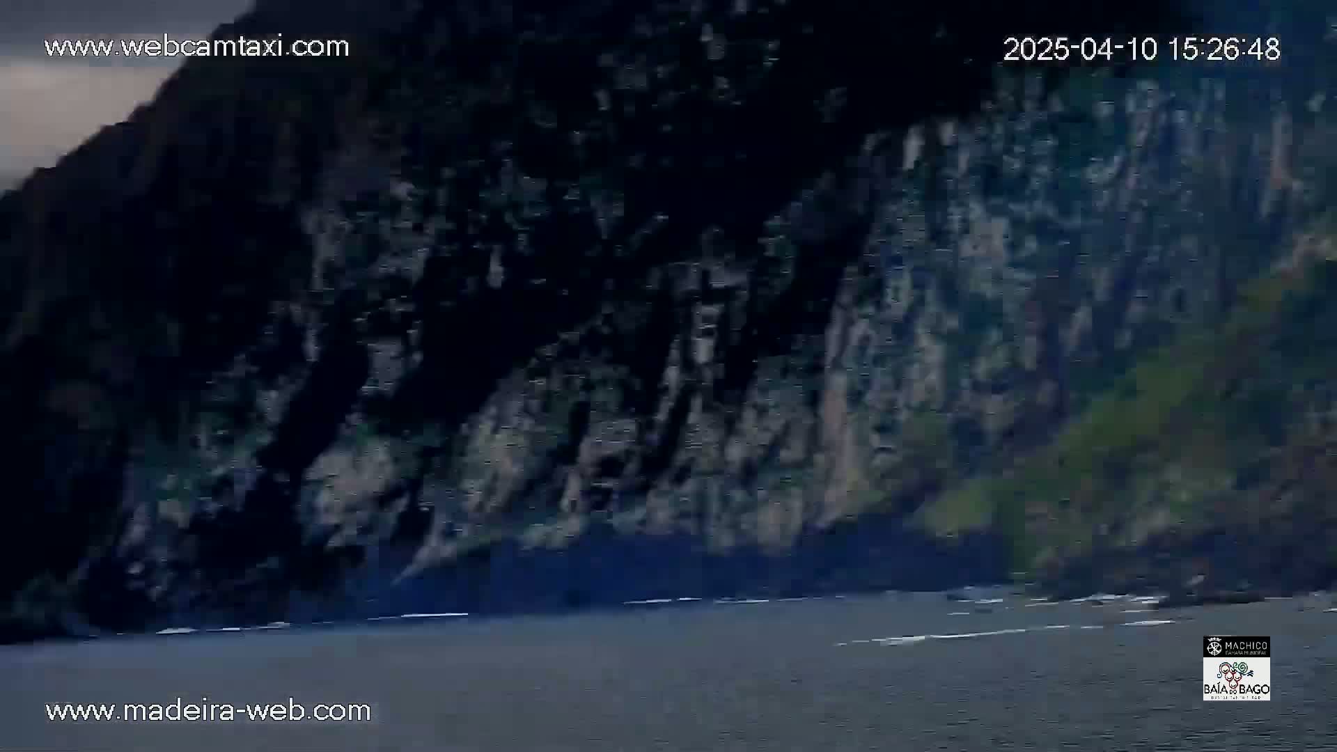 A dark, rocky cliff face rises sharply from calm, dark water under an overcast sky.