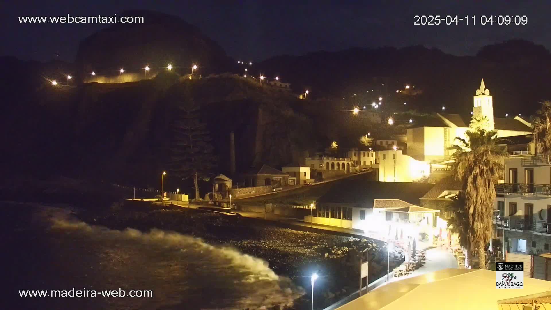 A nighttime coastal town scene with waves crashing on the shore, illuminated buildings climbing a hillside, and streetlights along a waterfront path.