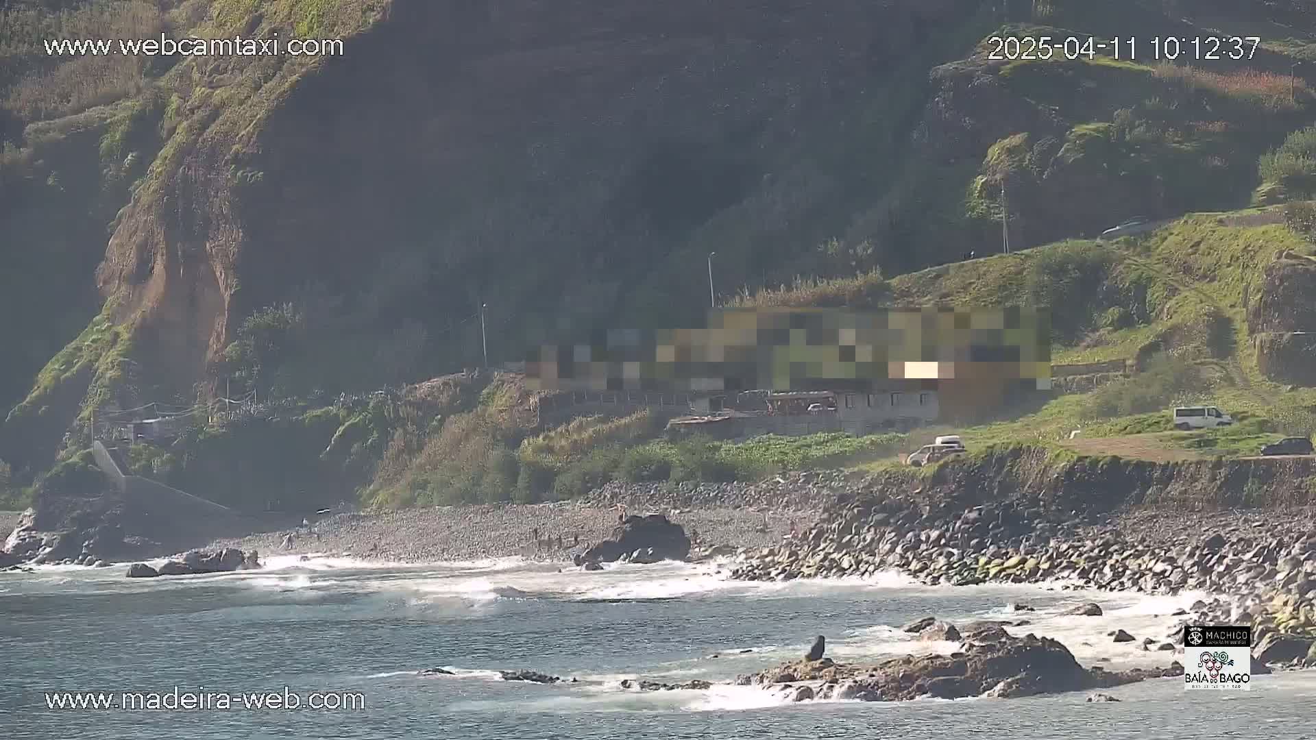 A rocky beach with some people on it is partially visible from a high vantage point, with small waves breaking near the shore and a hillside visible in the background under sunny conditions.