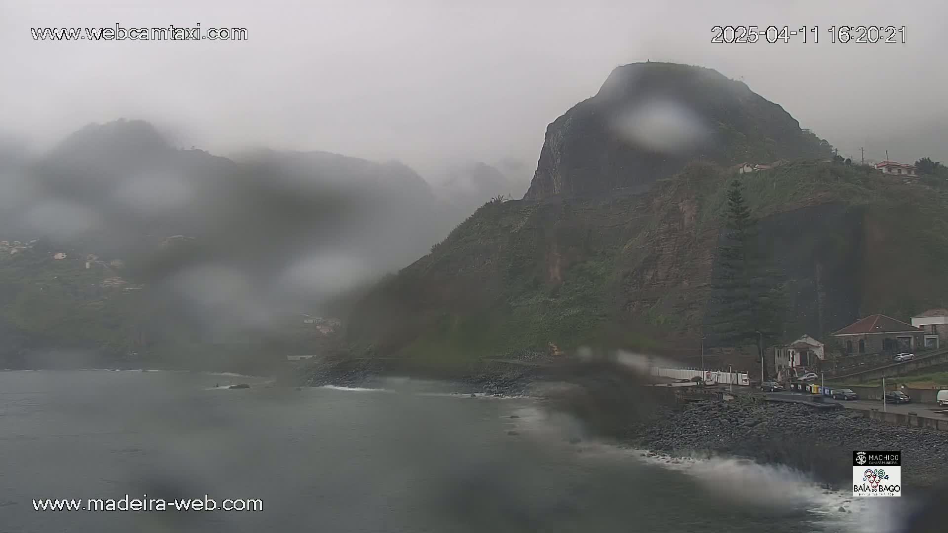 A rain-swept coastal scene shows dark, rocky cliffs and a gray ocean under an overcast sky.