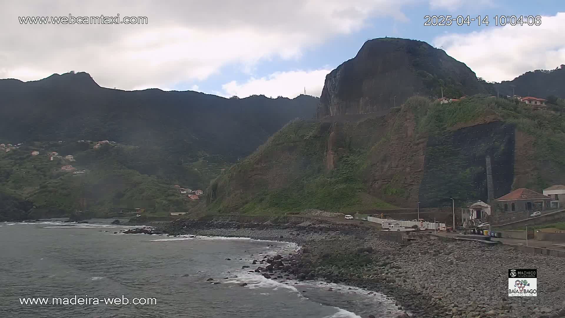 A rocky coastline with a steep, mountainous backdrop under partly cloudy skies.