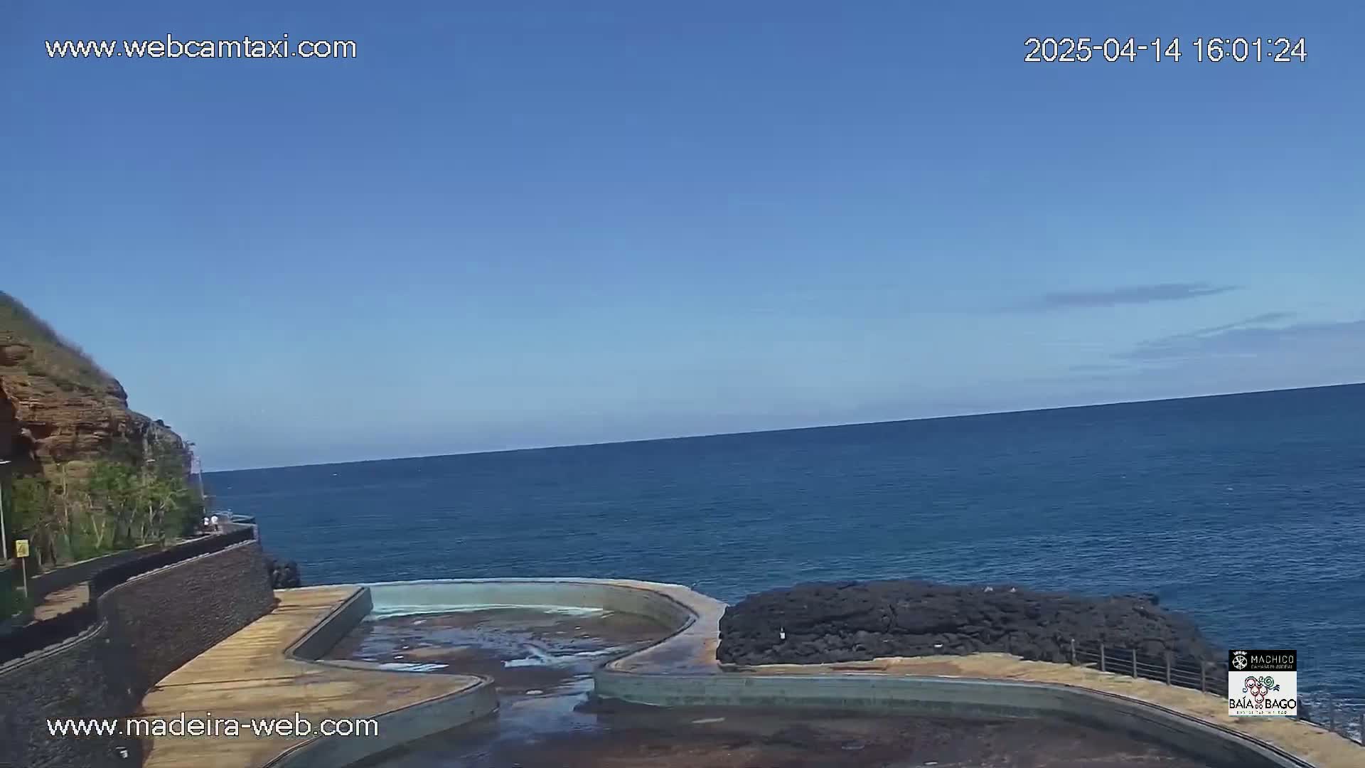 A coastal scene on a clear, sunny day shows a man-made pool next to a rocky cliff overlooking a calm ocean.