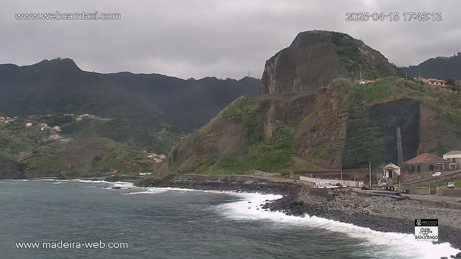 A rocky, dark-colored coastline with a dark, overcast sky features dark green vegetation growing on the cliffs, and small waves crash on a pebble beach.