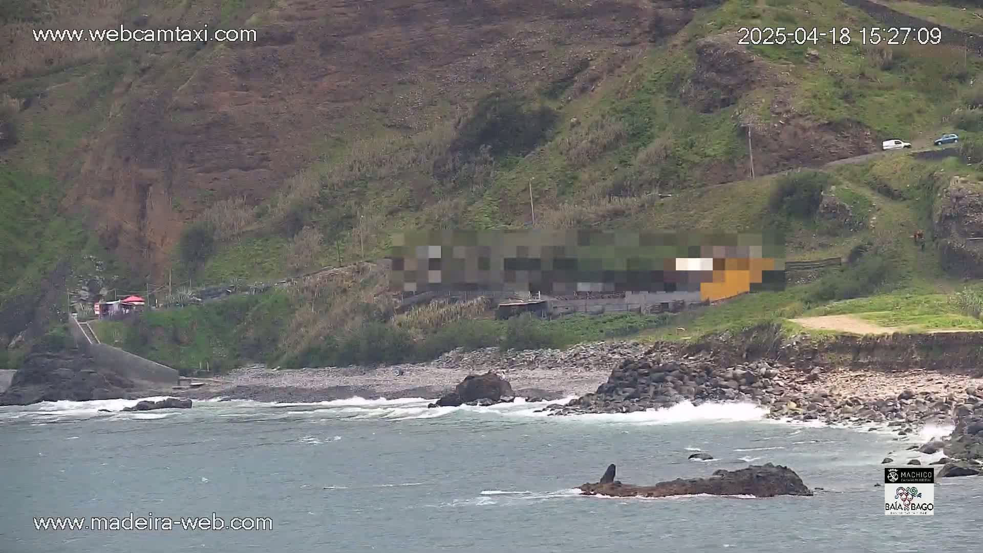 A rocky beach with small waves laps against the shore near a hillside with some buildings and sparse vegetation under overcast skies.