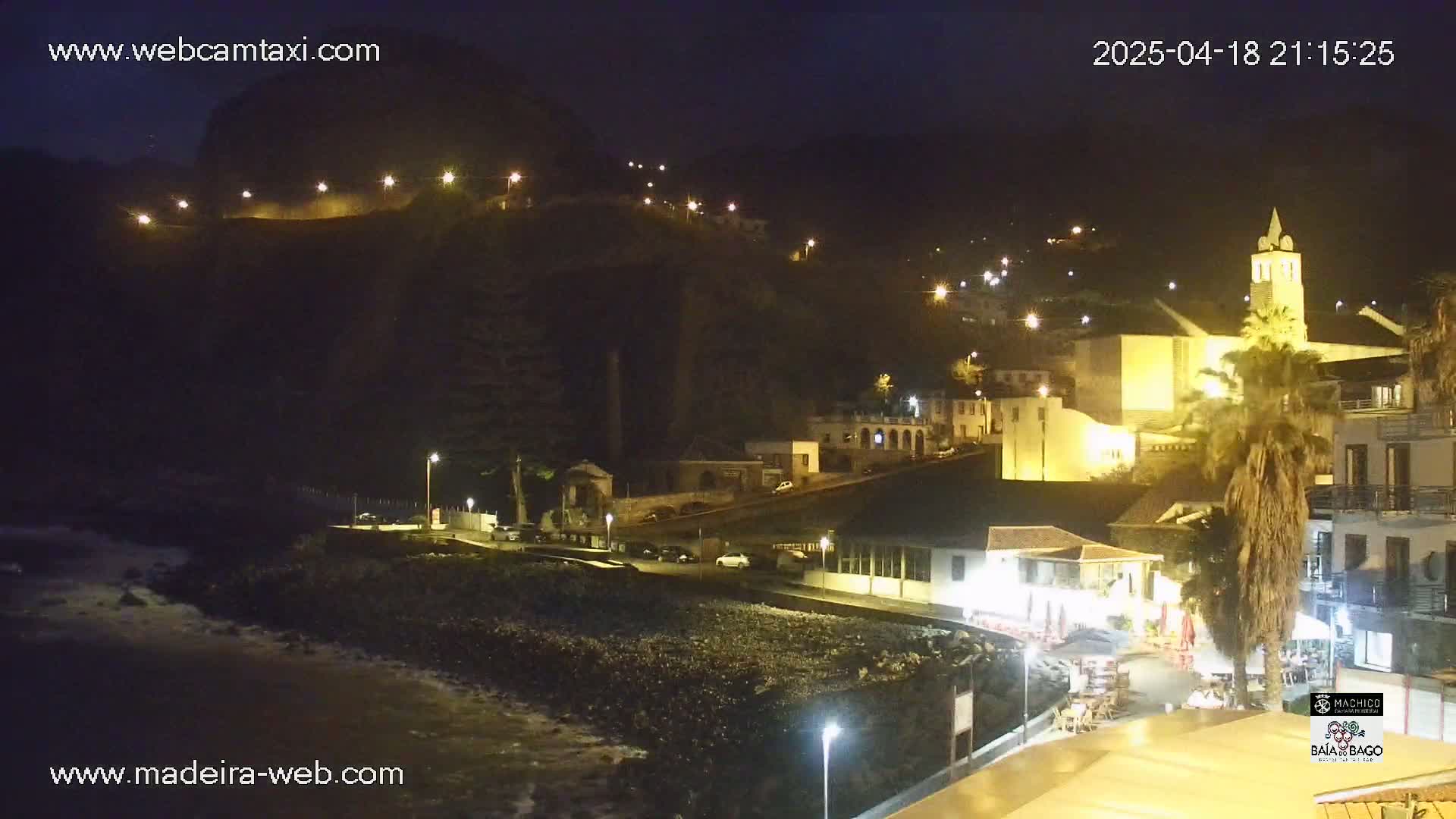 A nighttime coastal town scene shows buildings illuminated along a dark hillside, with a rocky beach and ocean visible in the foreground under a clear, dark sky.
