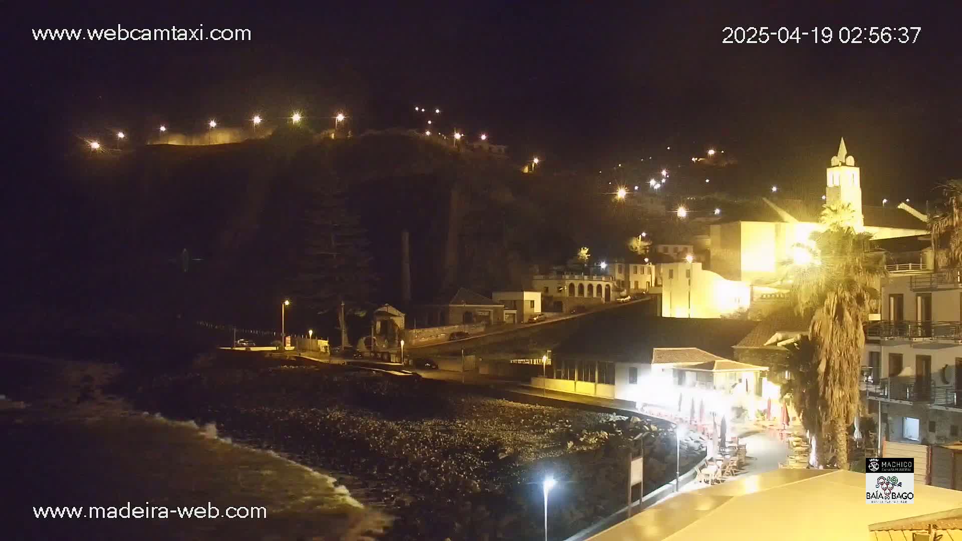 A nighttime coastal town is illuminated, with waves lapping at a dark, pebbled shore beneath a hill dotted with lights.