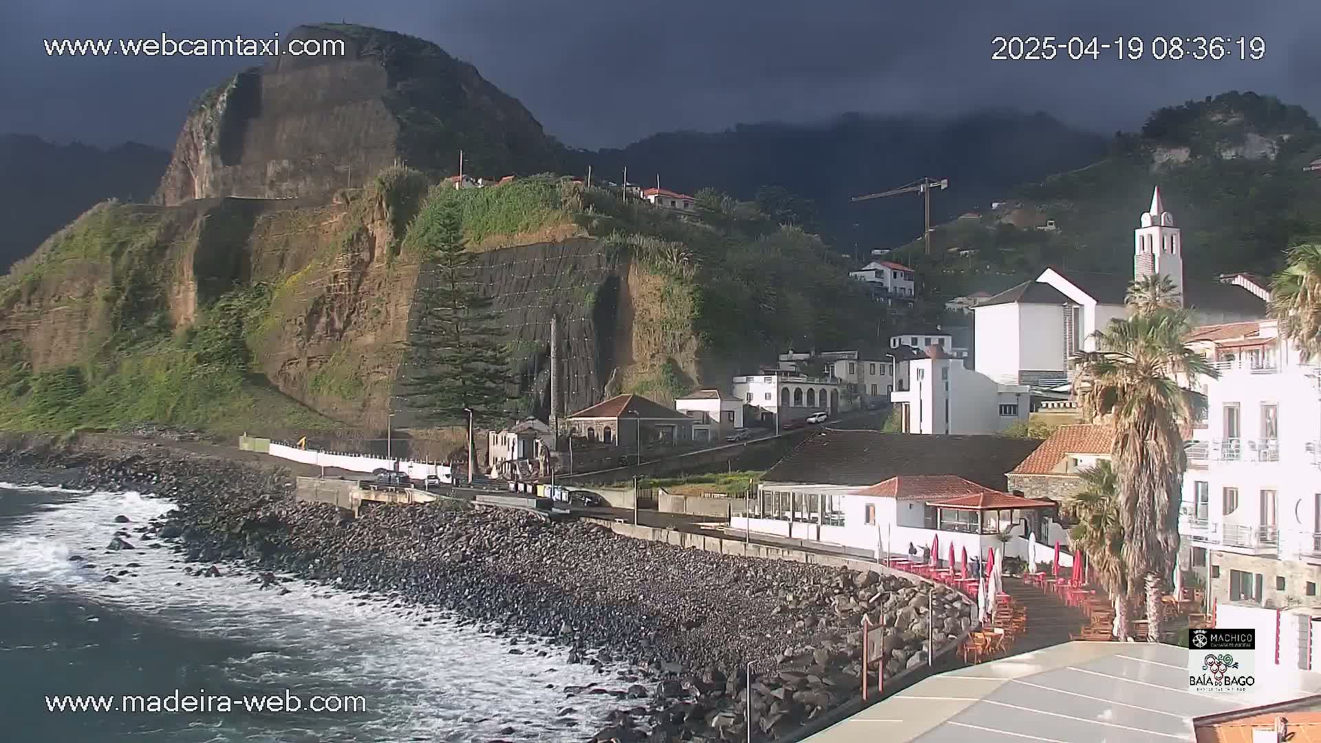 A coastal town nestled against a steep, dark cliff under an overcast sky, with small waves lapping a rocky shore.