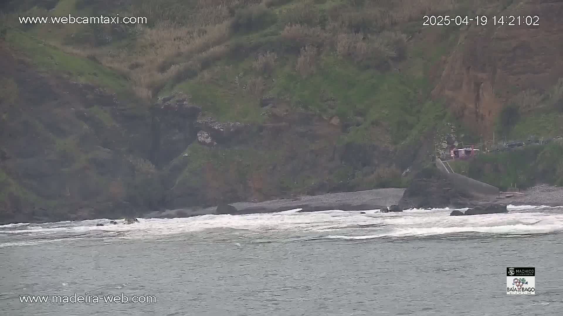 Ocean waves crash on a rocky shore near a green hillside.