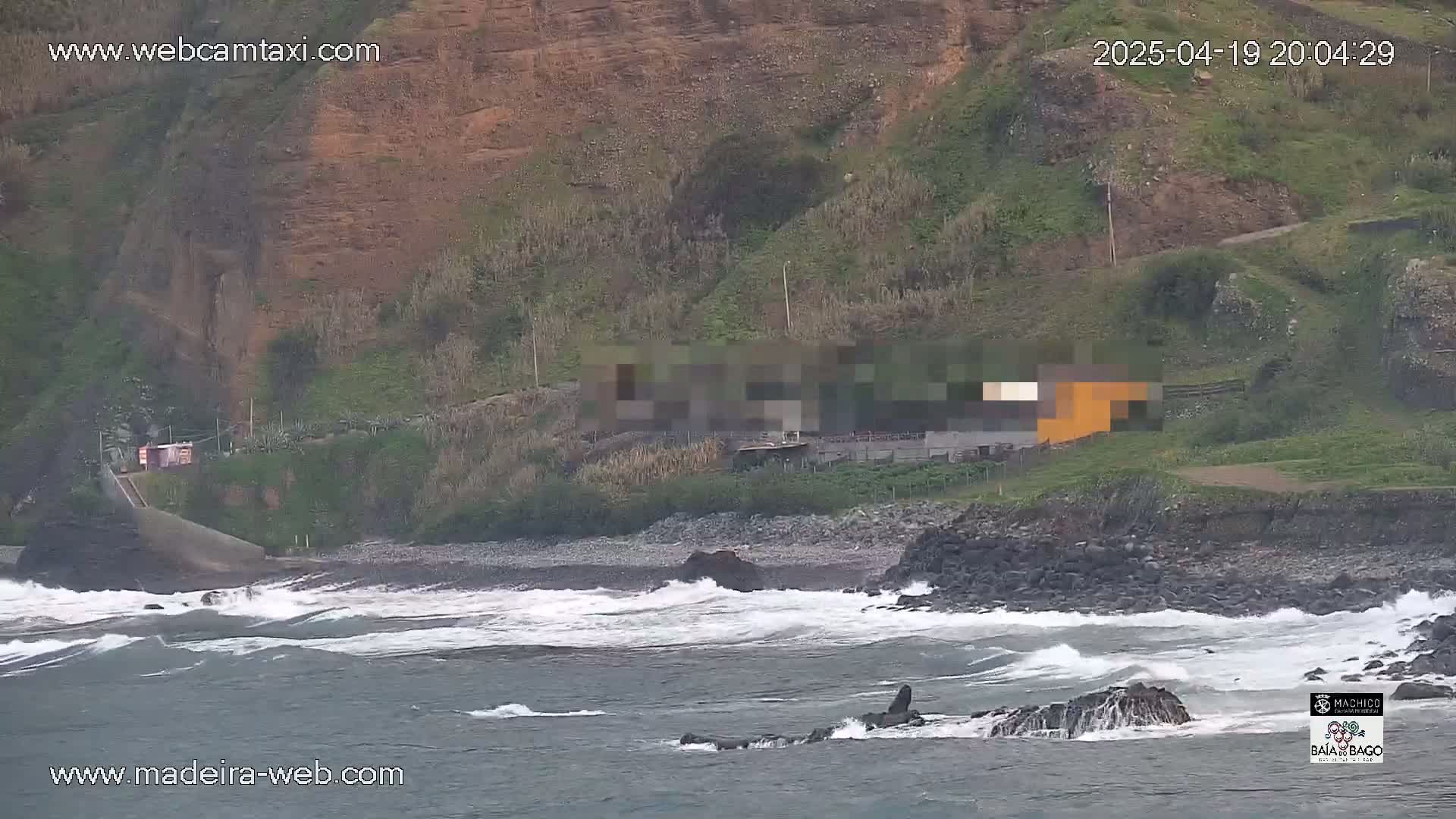 Waves crash on a rocky shore near a cliffside with sparse vegetation and a few buildings.