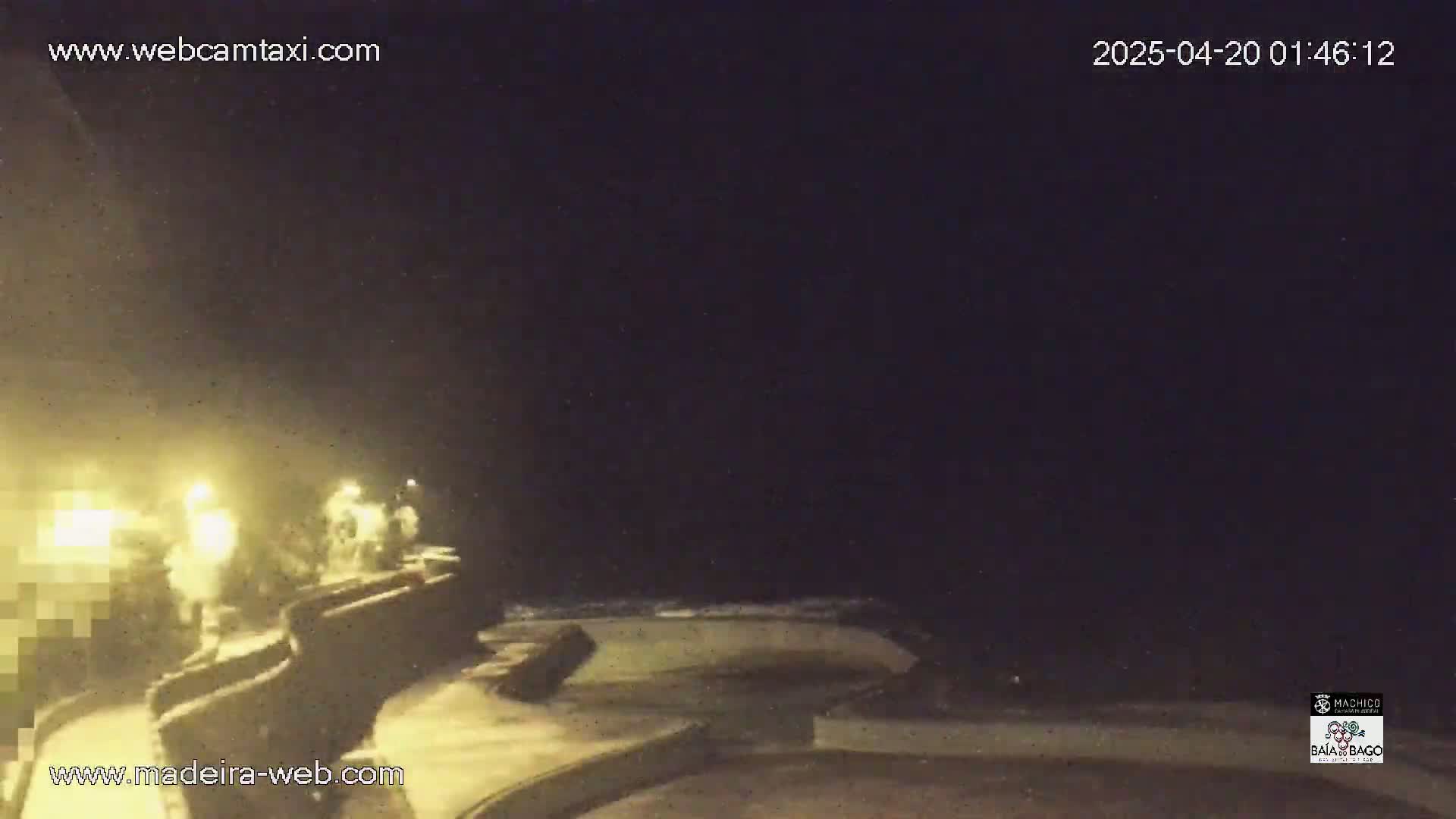 A nighttime, dimly lit coastal scene shows rough waves crashing against a stone wall, with some figures visible near the wall under artificial light.