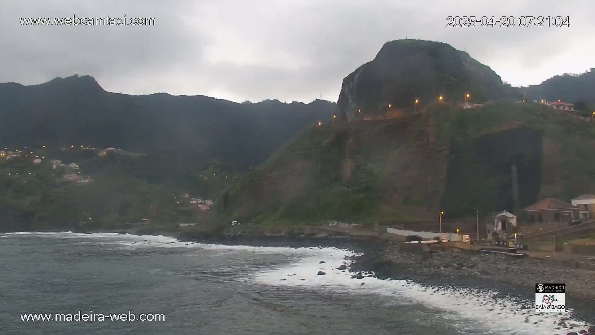 A dark, overcast day reveals a rocky coastline with dark green mountains in the background, small waves lapping at the shore, and a few buildings visible near the water.