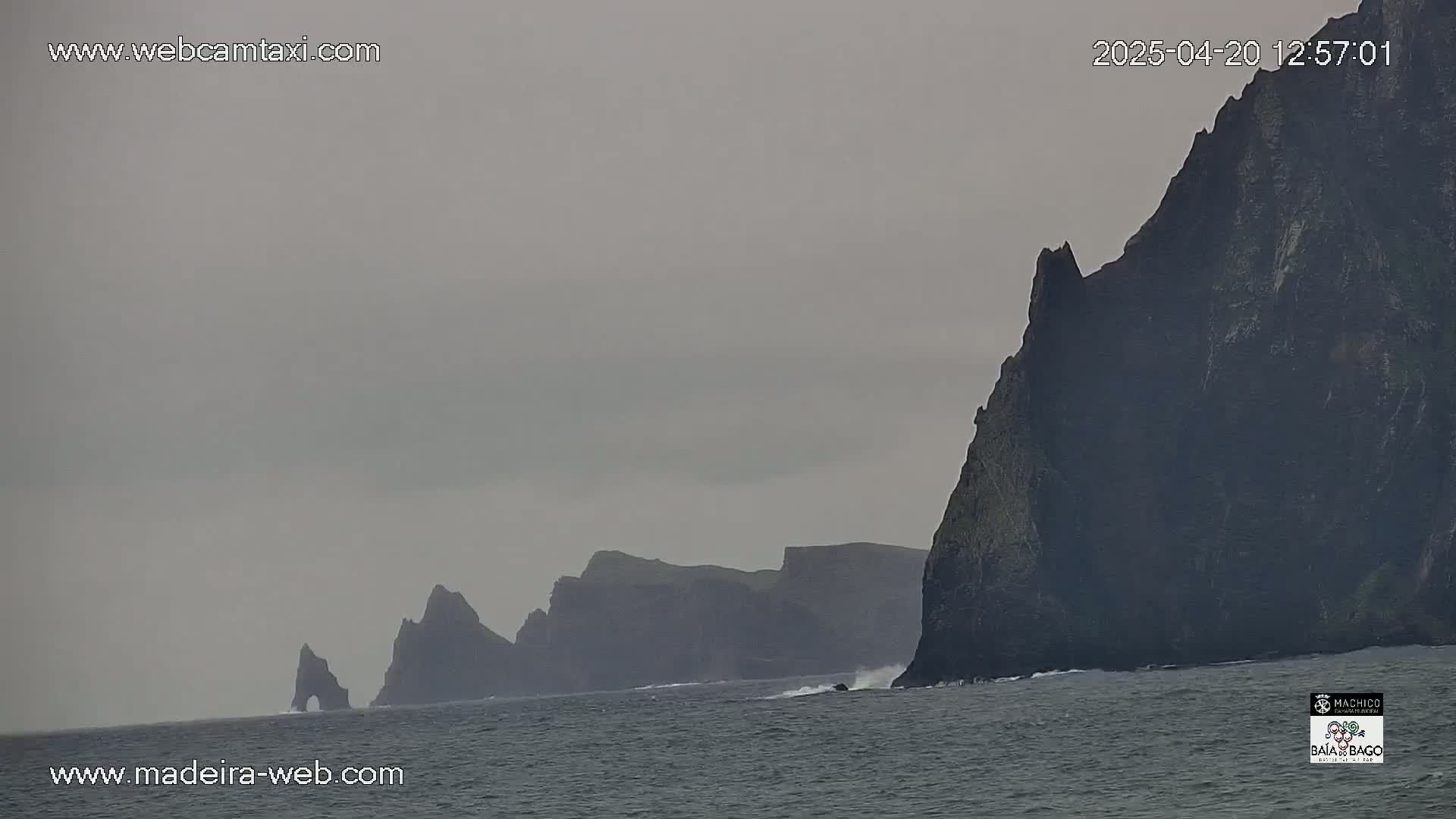 Dark, tall cliffs rise from a gray, choppy sea under an overcast sky.