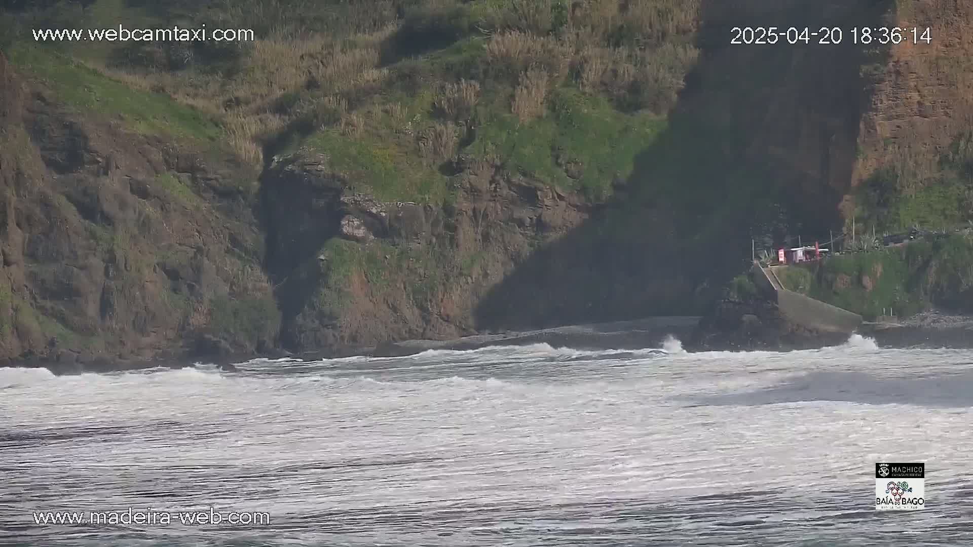 Ocean waves break near a rocky, vegetated cliff face on a partly sunny day.
