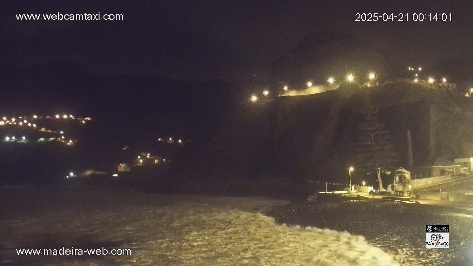 A nighttime coastal scene shows a dark, rocky cliff with lights illuminating a path and buildings, along with a dark, wavy ocean crashing on the shore below.