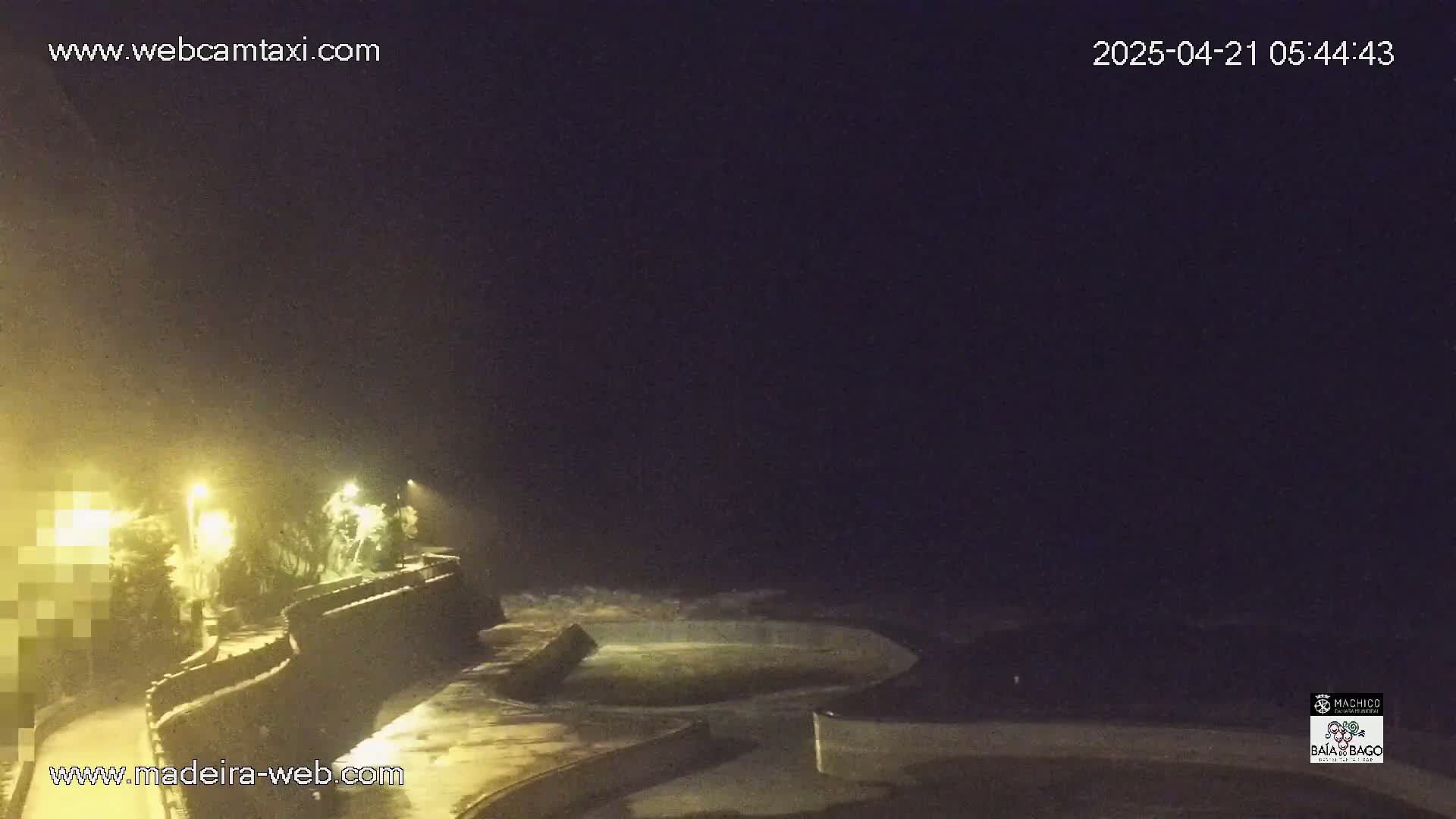 A nighttime, dimly lit coastal scene shows a dark ocean with waves crashing against a seawall; some lights illuminate a walkway along the wall.