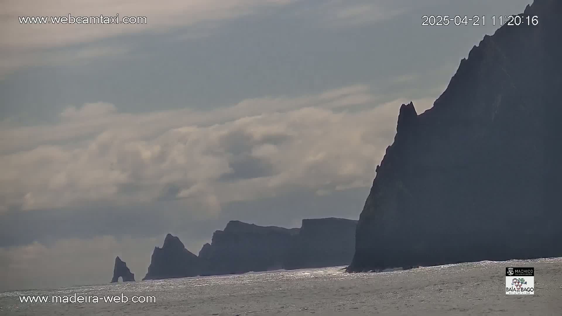 Dark, rocky cliffs rise from a calm ocean under a cloudy sky.