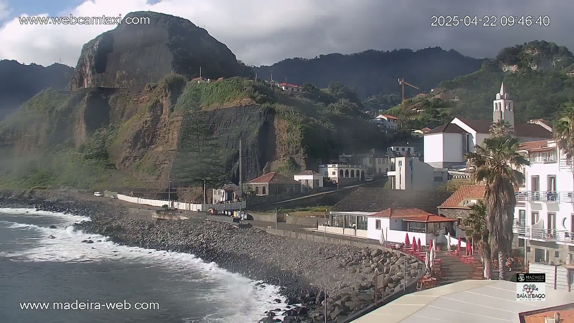 Ocean waves crash against a rocky shoreline, next to a village nestled against a steep, partially-reinforced cliffside beneath cloudy skies.