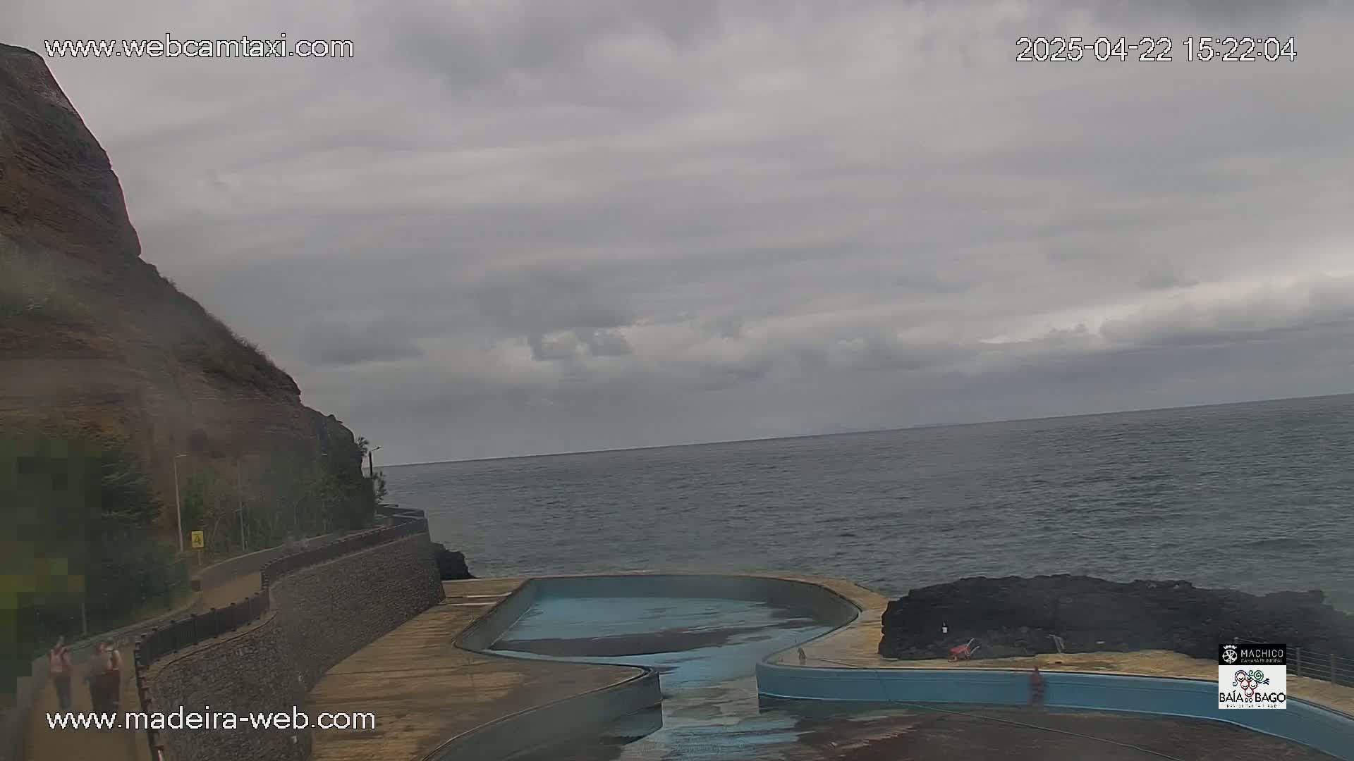 An outdoor view shows an empty, curved, light-blue pool next to a dark-colored, rocky cliff overlooking a dark-grey ocean under an overcast sky.