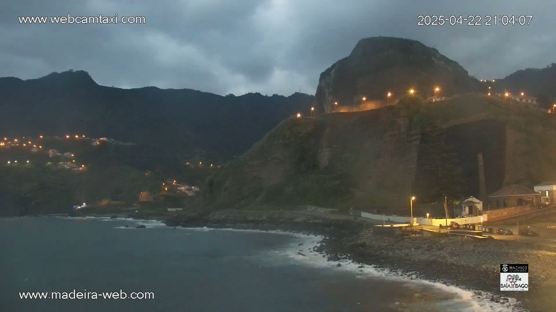 A dark, overcast night scene shows a rocky coastline with a village nestled into the hills, illuminated by streetlights, under a dark and cloudy sky.