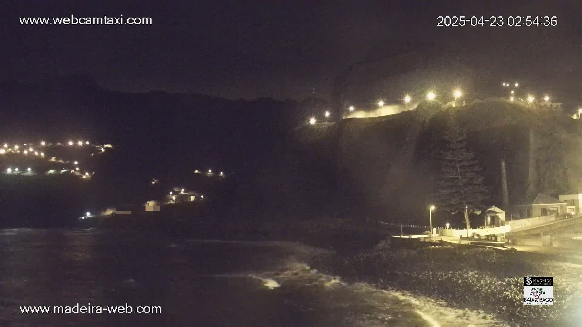 A nighttime coastal scene shows a dark, rocky shoreline with waves, illuminated buildings on a hillside, and lights along a roadway.