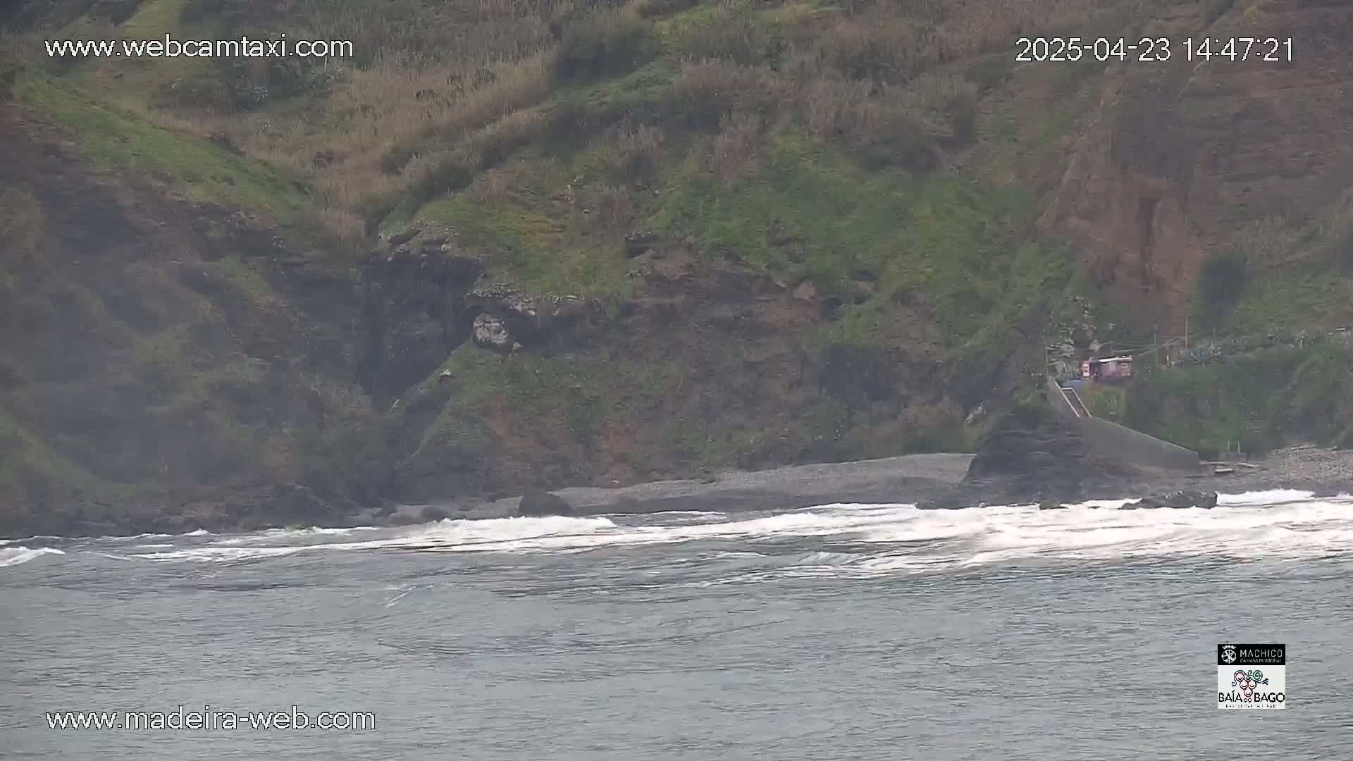 A dark-colored, rocky beach with small waves laps against a shore partially obscured by an overcast sky.