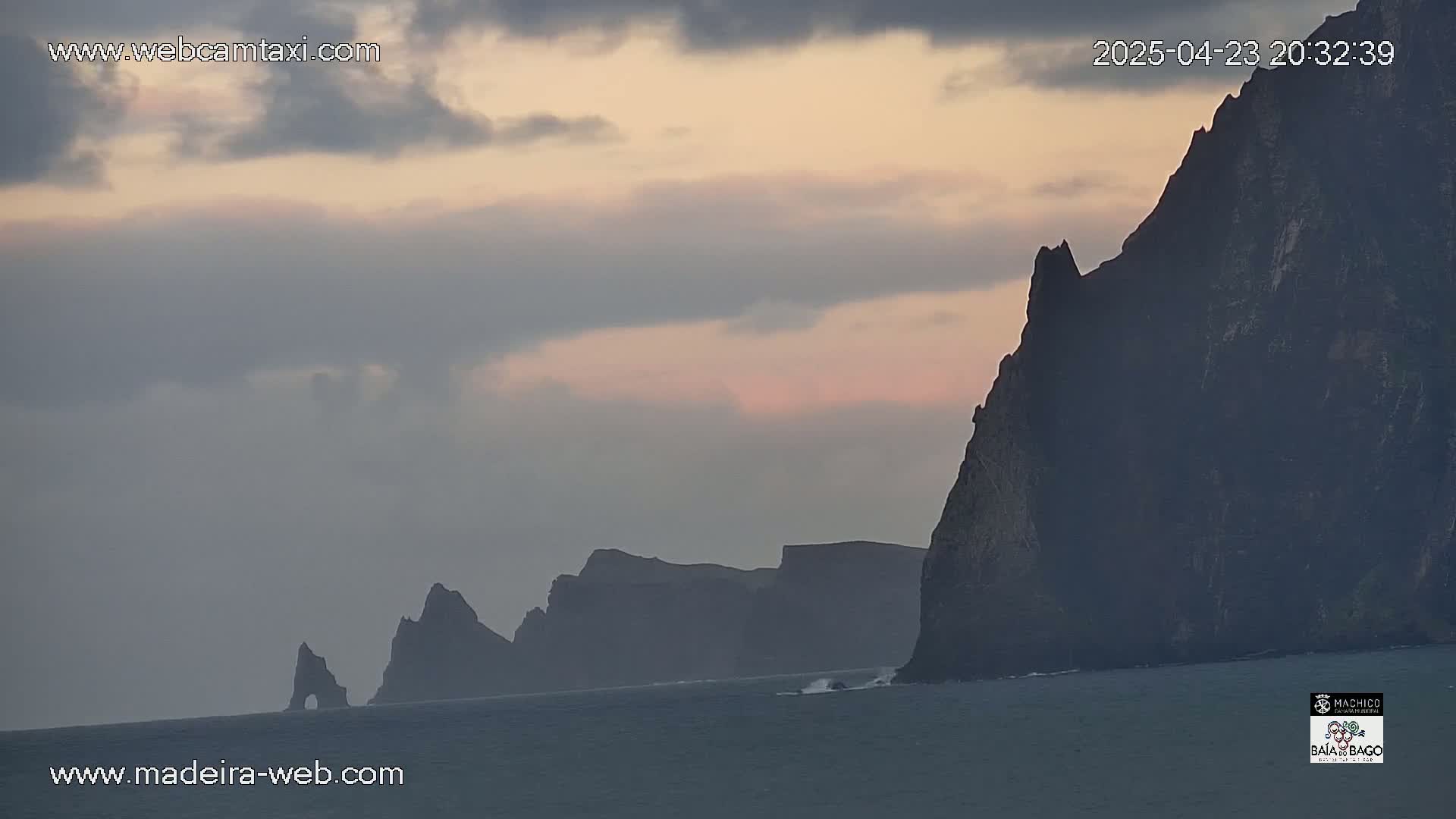 Dark, rocky cliffs rise from a calm ocean under a pale, cloudy sunset sky.