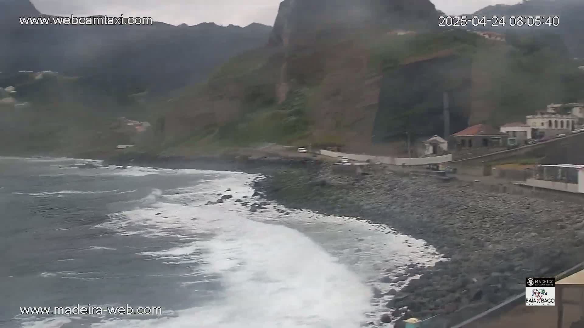 Waves crash on a rocky shoreline next to a road and buildings at the base of a cliff under an overcast sky.