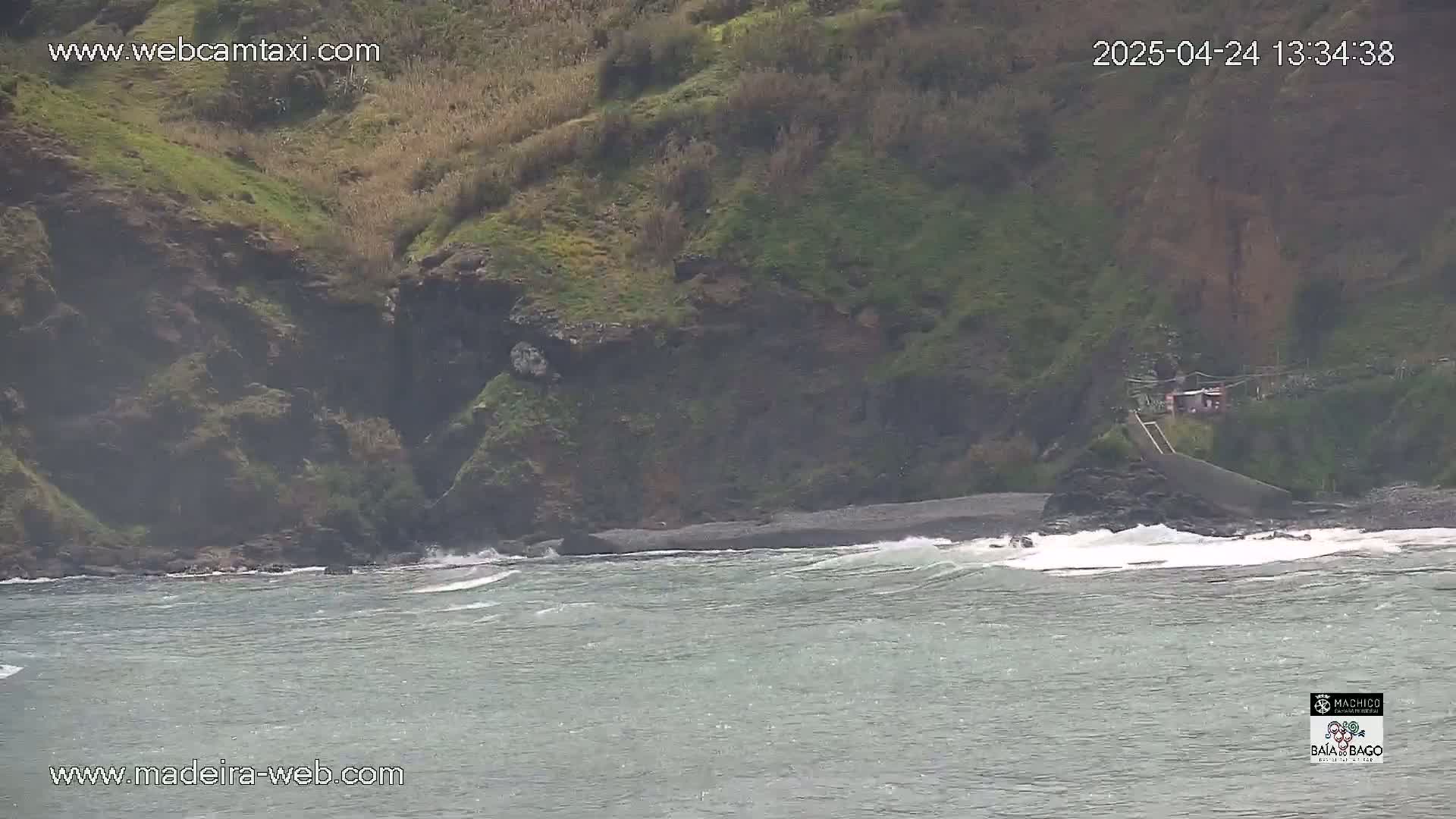 A rocky, green hillside descends to a dark-sand beach where small waves lap the shore in overcast conditions.
