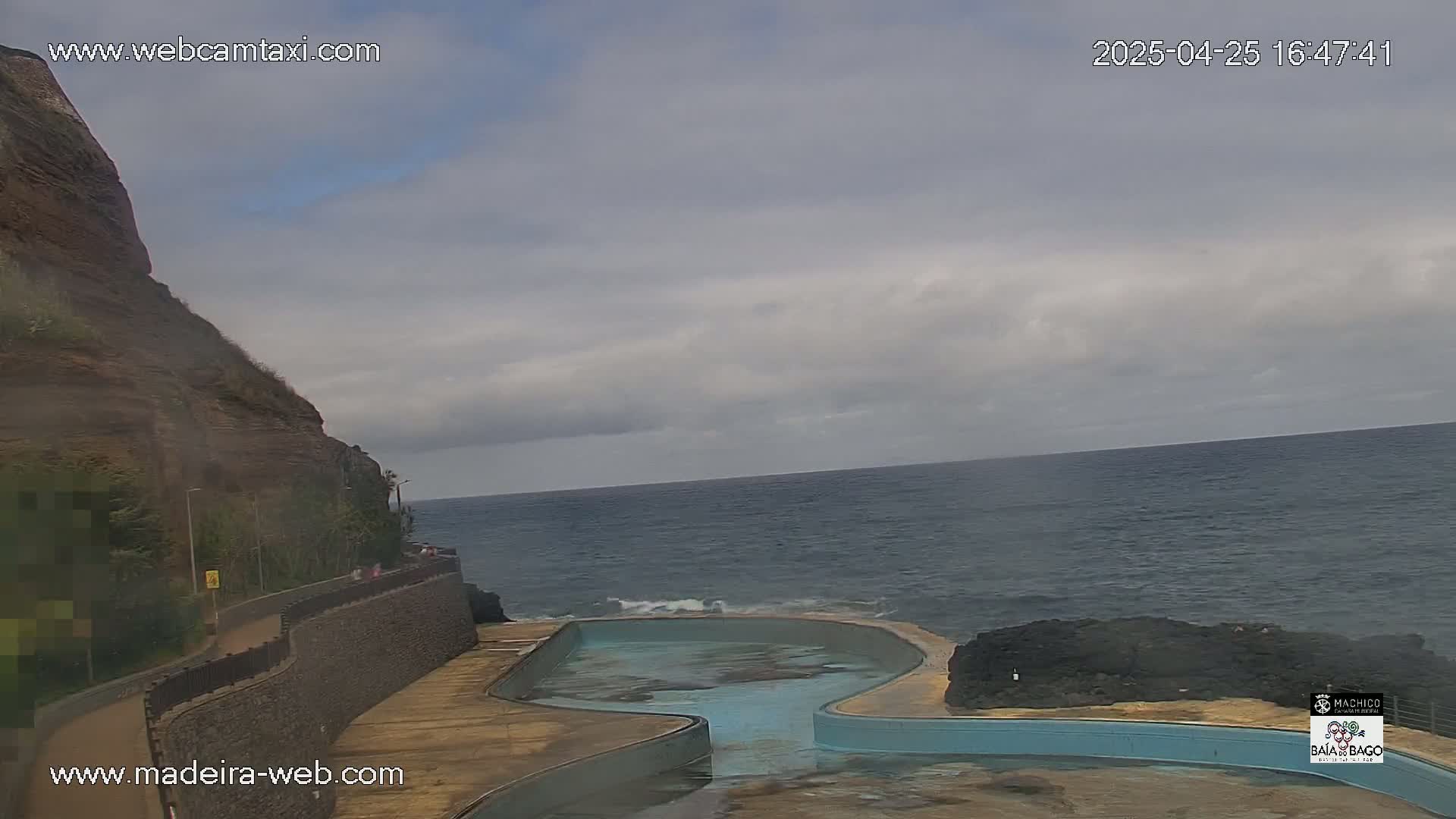 A coastal scene shows an ocean with a partly cloudy sky above a curved, empty pool built into a rocky shore next to a cliff.