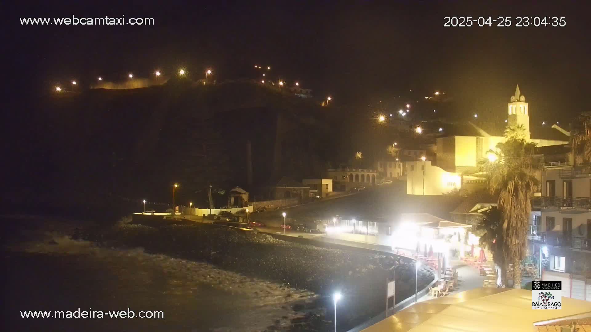 A nighttime coastal town scene shows a dark, rocky beach with waves, illuminated buildings on a hillside, and streetlights along a waterfront walkway.