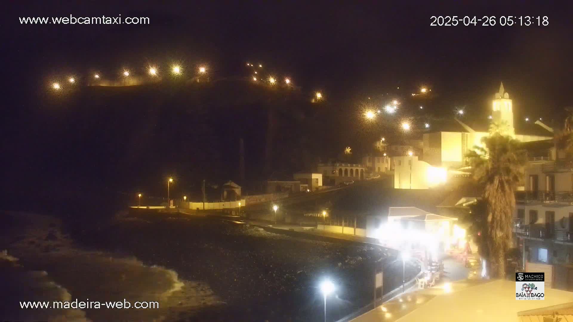 A nighttime coastal town scene shows a dark, rocky beach with small waves, illuminated buildings on a hillside, and a tall church steeple.
