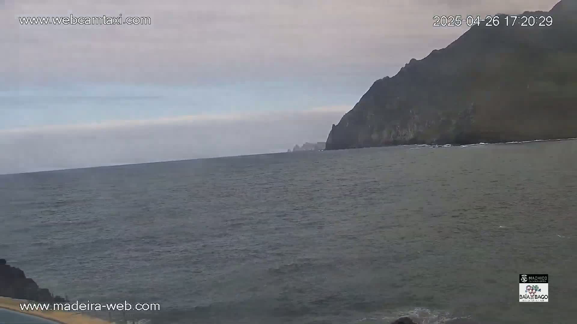 A dark, overcast sky hangs over a calm, gray ocean meeting a rocky, dark-colored cliff.