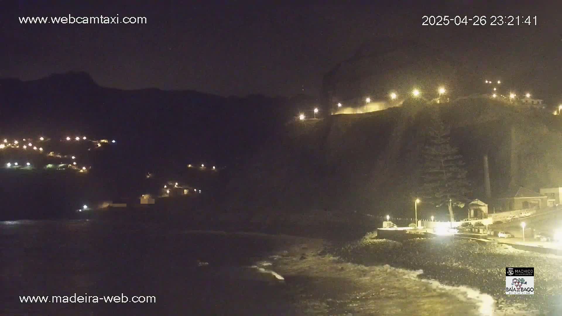A nighttime coastal scene shows a dark cliff illuminated by lights, with a small town nestled on its slopes, and gentle waves lapping the shore.