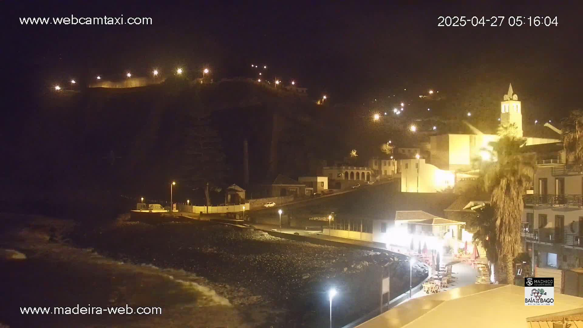 A nighttime coastal town with illuminated buildings and a rocky beach, lit by streetlights and the glow from businesses, with gentle waves lapping the shore.