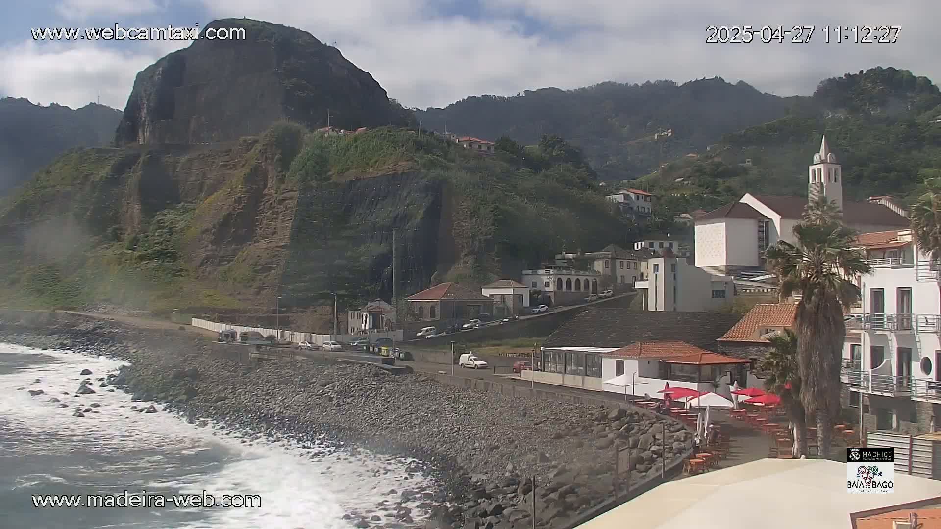 Waves crash on a rocky shore next to a village nestled against a steep, green mountain under a partly cloudy sky.
