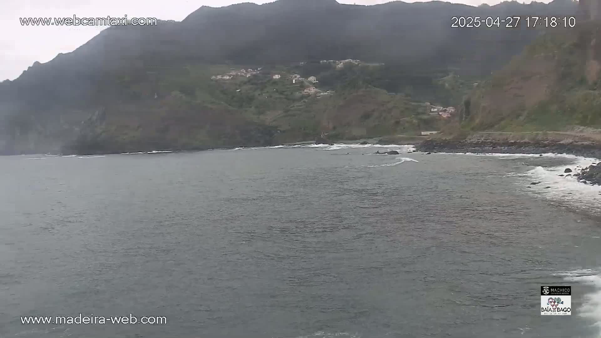 A gray, overcast day reveals a calm ocean bay curving around a rocky shoreline, with a small village nestled into the green hills beyond.