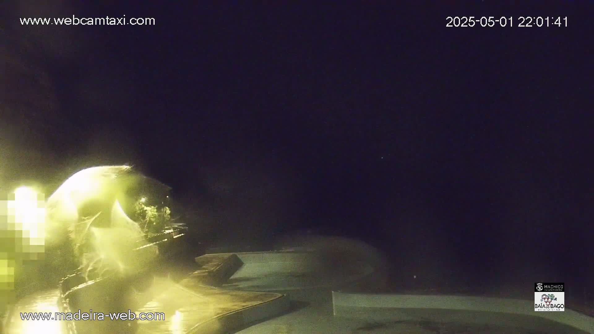A nighttime view shows a brightly lit area of water with splashing, possibly a fountain or wave action, under a dark sky.