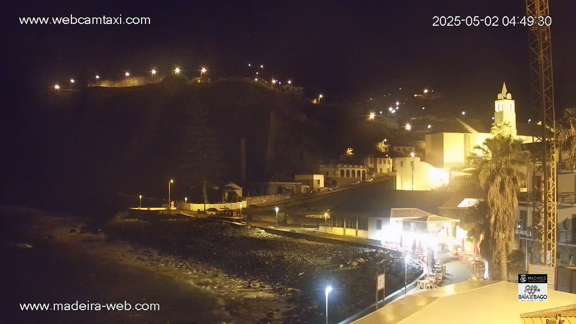A nighttime coastal town scene with a rocky beach and gently lapping waves, illuminated by streetlights and building lights on a hillside.