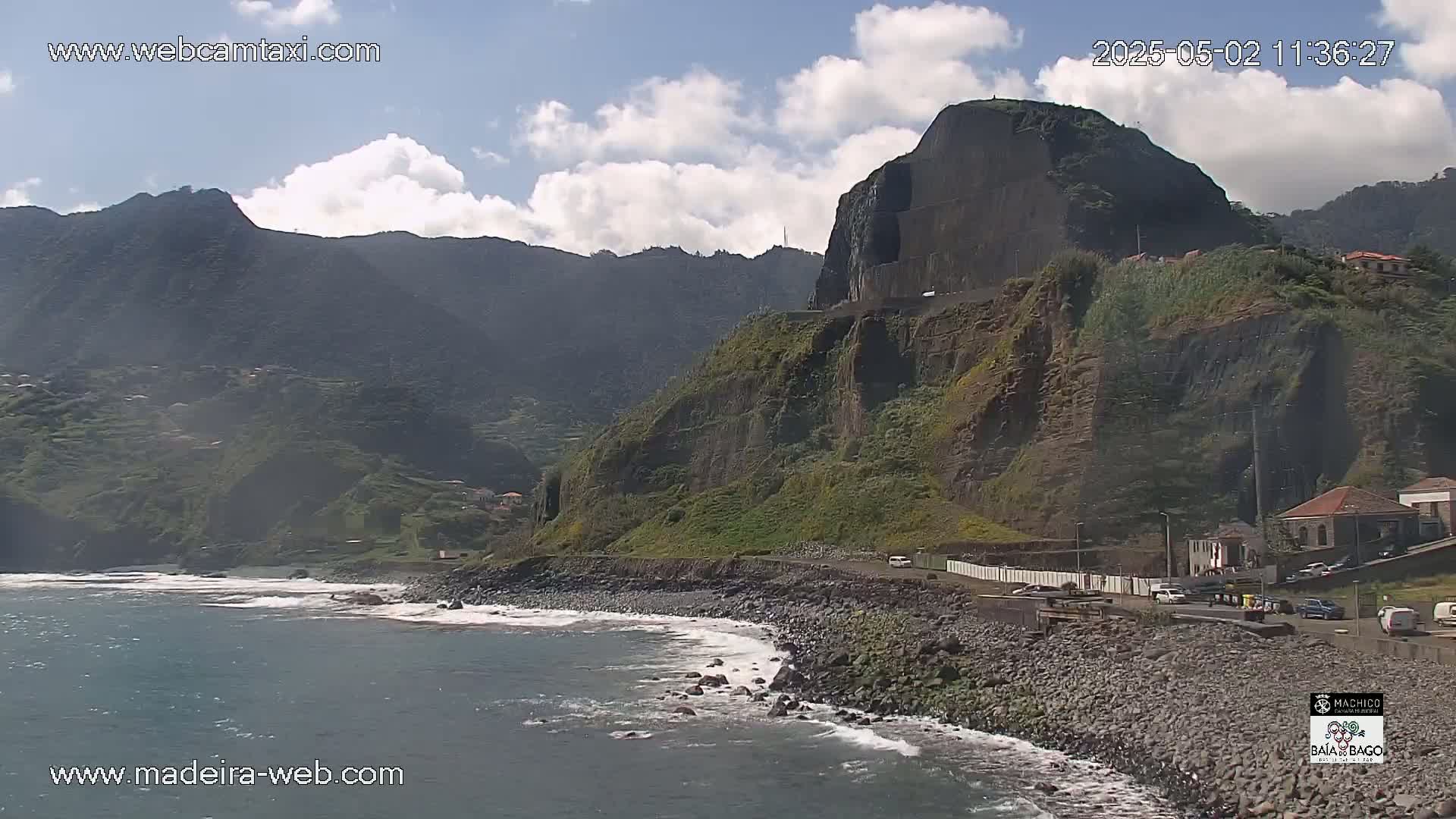 A rocky coastline with a steep, dark cliff face rising behind a pebble beach and a calm ocean under a partly cloudy sky.