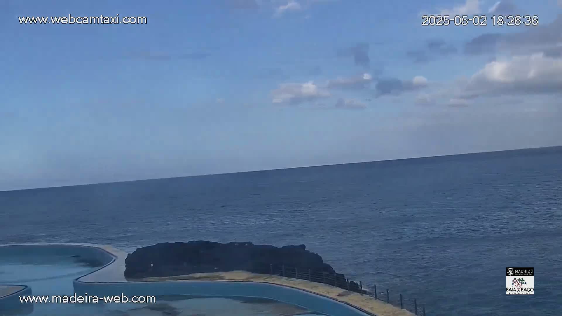 A mostly clear sky overlooks a calm ocean next to a dark, rocky shoreline with a light blue pool.