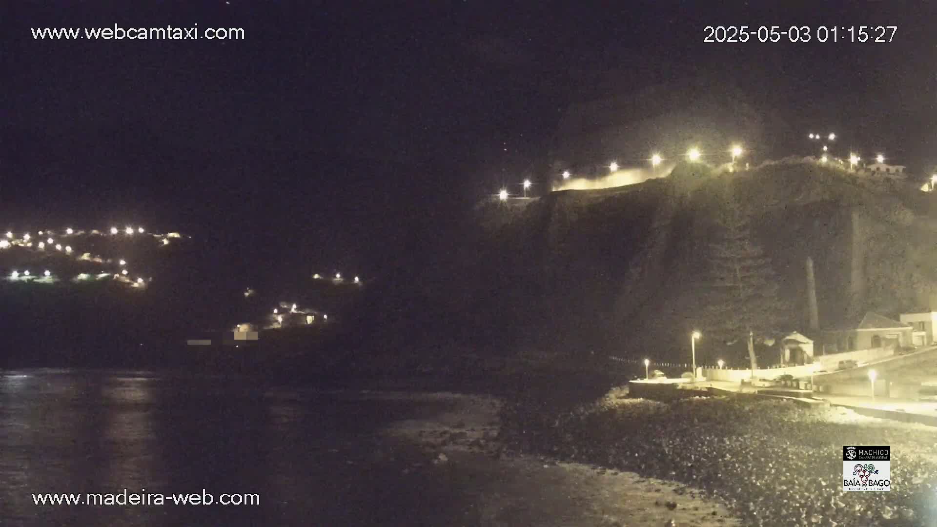 A nighttime coastal scene shows a dark, rocky cliff illuminated by lights, with a calm body of water below and small settlements visible on the hillsides.