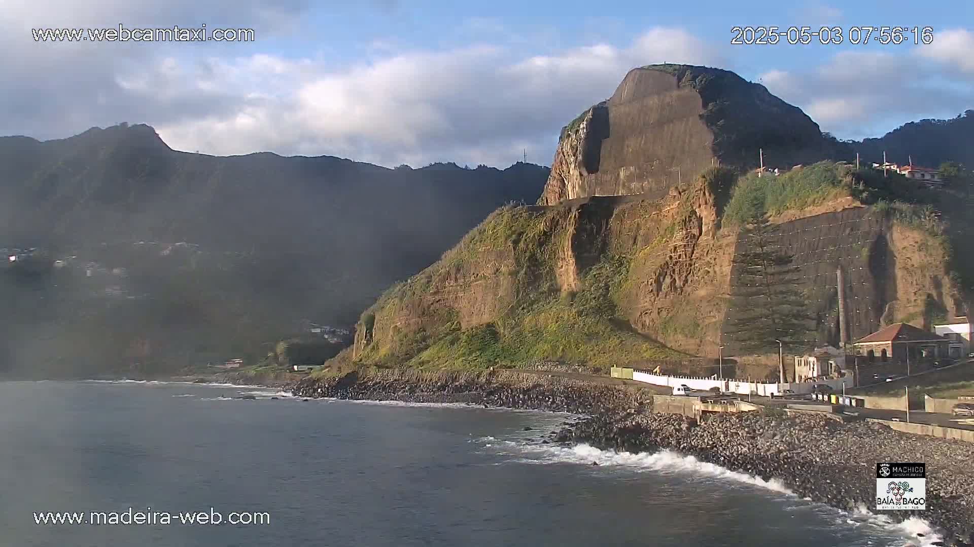 A rocky coastline with a steep cliff face and sparse vegetation overlooks a calm ocean under a partly cloudy sky.