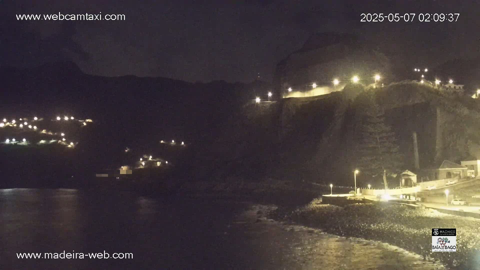 A nighttime coastal scene shows a dark cliff face illuminated by lights, with a small town nestled below and dark water lapping at the shore.