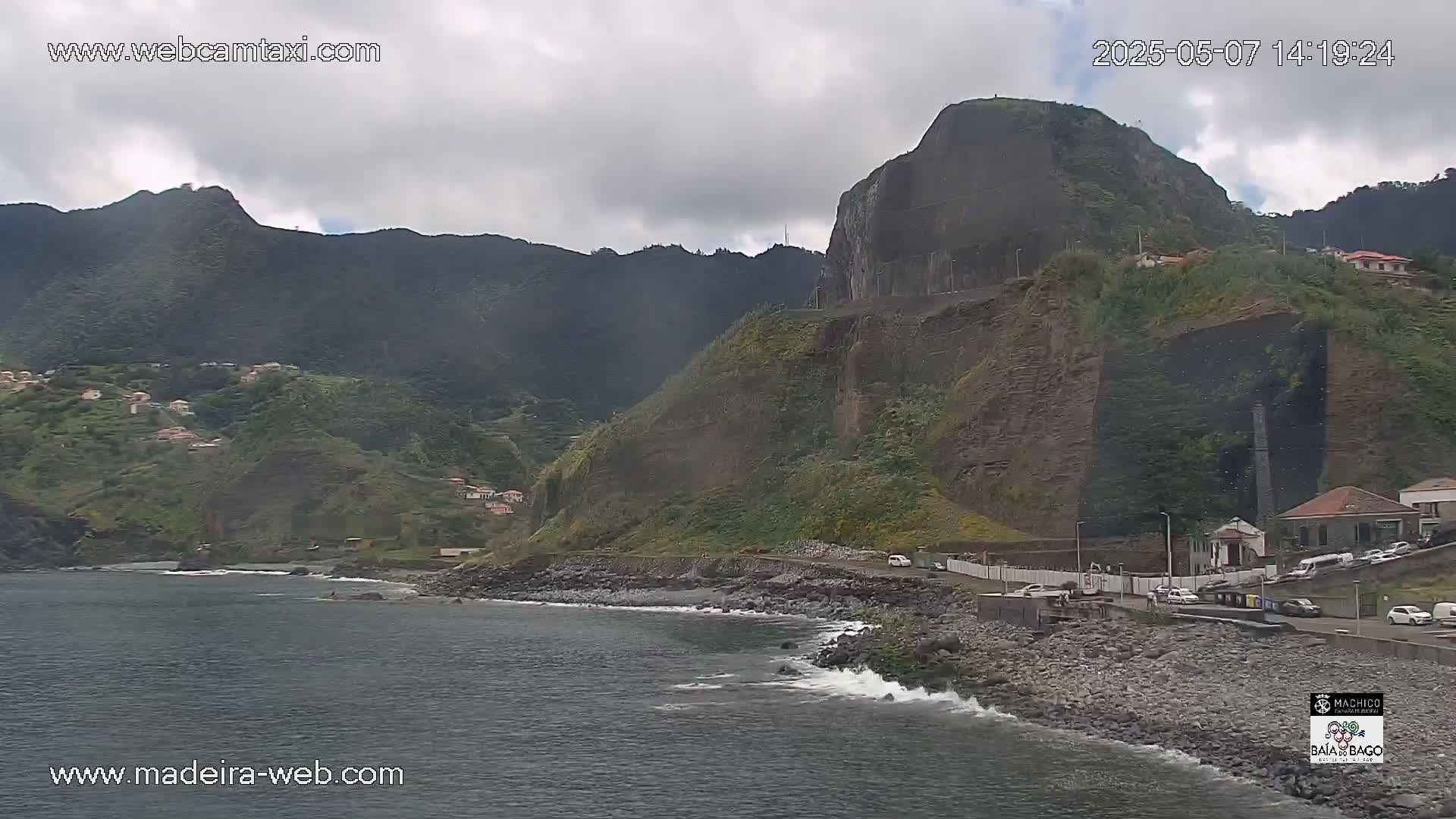 A rocky coastline with a dark, overcast sky features a village nestled into the hillside, with a steep cliff and calm ocean water.