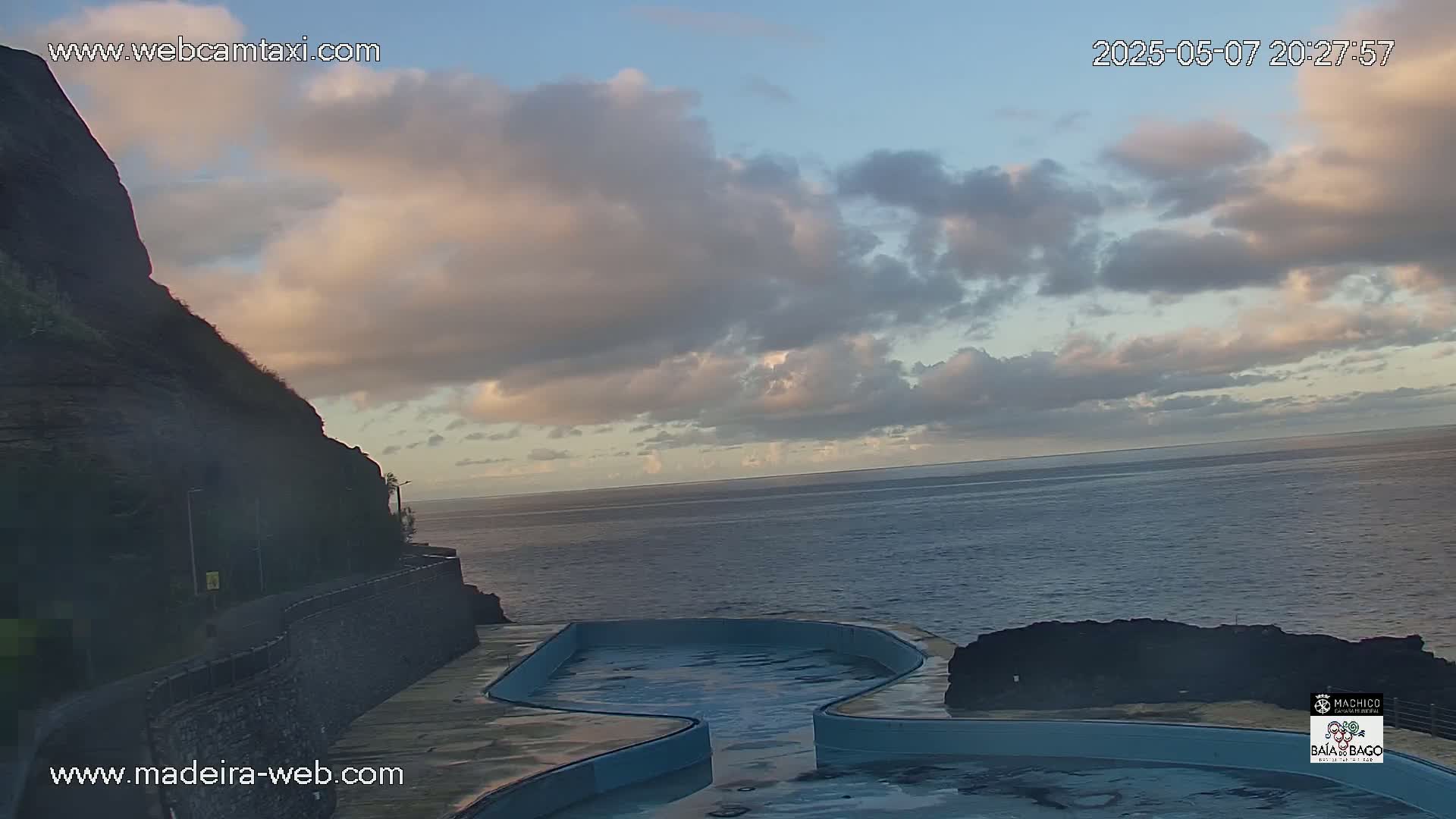 A partially cloudy sky overlooks a dark-colored cliff and ocean, with a light-blue oceanside pool in the foreground.